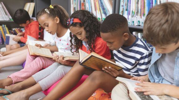 A group of children sitting next to each other read books on the floor with a bookshelf behind them.